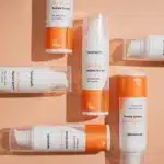 Verschillende flesjes Hemptouch - Skin Perfection - CBD Azelaic Serum voor gezicht (30ml) met witte en oranje verpakking staan op een perzikkleurig oppervlak, van bovenaf gezien. Productgegevens zijn zichtbaar op de etiketten.