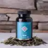 Een flesje MindScopic - CLEAR FOCUS™ Cafeïnevrije Capsules (50 stuks) rust op een wit oppervlak met losse kruidenbladeren in de buurt en een stenen muur op de achtergrond.