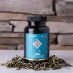 Een flesje MindScopic - CLEAR FOCUS™ Cafeïnevrije Capsules (50 stuks) rust op een wit oppervlak met losse kruidenbladeren in de buurt en een stenen muur op de achtergrond.