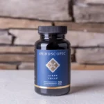 Een flesje MindScopic - CLEAR FOCUS™ Capsules (50 stuks) staat op een licht oppervlak voor een stenen muur.