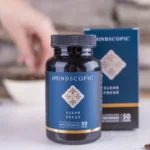 Een fles MindScopic - CLEAR FOCUS™ Capsules (50 stuks) wordt getoond voor de doos, met op de achtergrond een hand die naar een kom reikt en koffiebonen verspreid op het oppervlak.