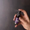 Een hand spuit een klein flesje met de tekst "MindScopic - SLEEPWELL SPRAY met melatonine (15ml)" tegen een donkere achtergrond.