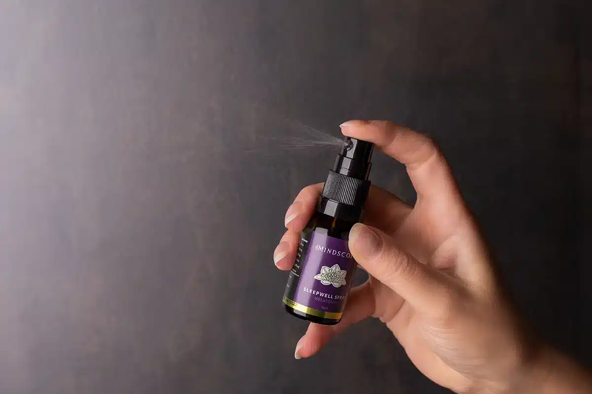 Een hand spuit een klein flesje met de tekst "MindScopic - SLEEPWELL SPRAY met melatonine (15ml)" tegen een donkere achtergrond.