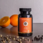 Een fles MindScopic - MOODMASTER™ capsules (50 stuks) staat op het aanrecht met een sinaasappel, een sinaasappelhelft en verspreide peperkorrels en gedroogde kruiden.