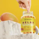 Een hand pakt een gummie uit een potje met het label "Harmony - CBD / CBG Relax Gummies (60 stuks) - THT datum 21-11-2025" op een marmeren oppervlak, met een mango en gele achtergrond.