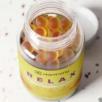 Een geopend flesje Harmony - CBD / CBG Relax Gummies (60 stuks) - THT datum 21-11-2025 laat gele en oranje gummies zien, met het deksel ernaast op een gespikkeld oppervlak.