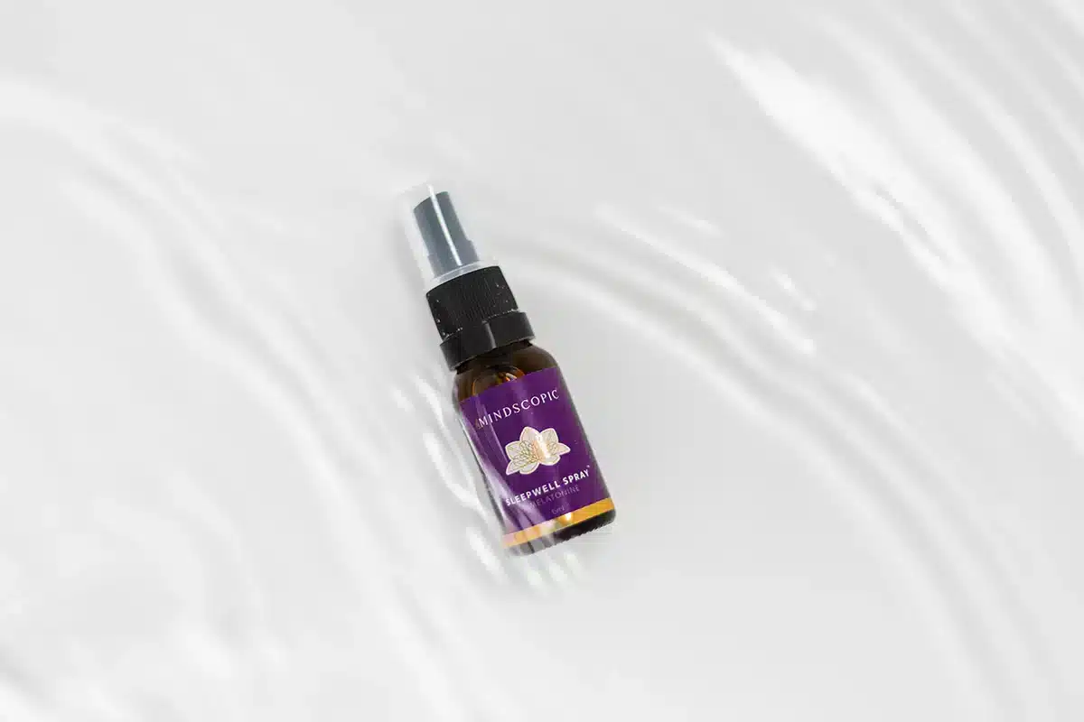 Een klein bruin sprayflesje met de tekst "MindScopic - SLEEPWELL SPRAY met melatonine (15ml)" met een paars en gouden ontwerp staat op een wit, waterig oppervlak.