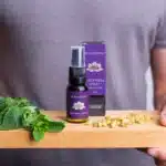 Een persoon houdt een houten plank vast met verse munt, kamillebloemen en een MindScopic - SLEEPWELL SPRAY met melatonine (15ml) flesje en doosje.