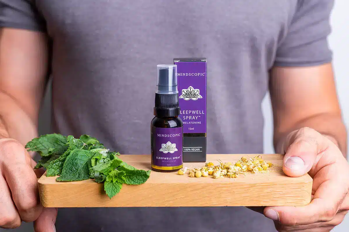 Een persoon houdt een houten plank vast met verse munt, kamillebloemen en een MindScopic - SLEEPWELL SPRAY met melatonine (15ml) flesje en doosje.